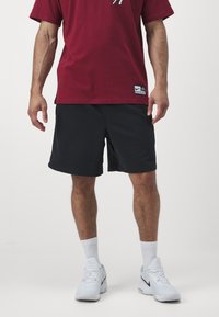 T-shirt vermelha de manga curta com logotipo, combinada com calções pretos e sapatilhas brancas, apresentando meias brancas até ao tornozelo. Design simples, ajuste casual.