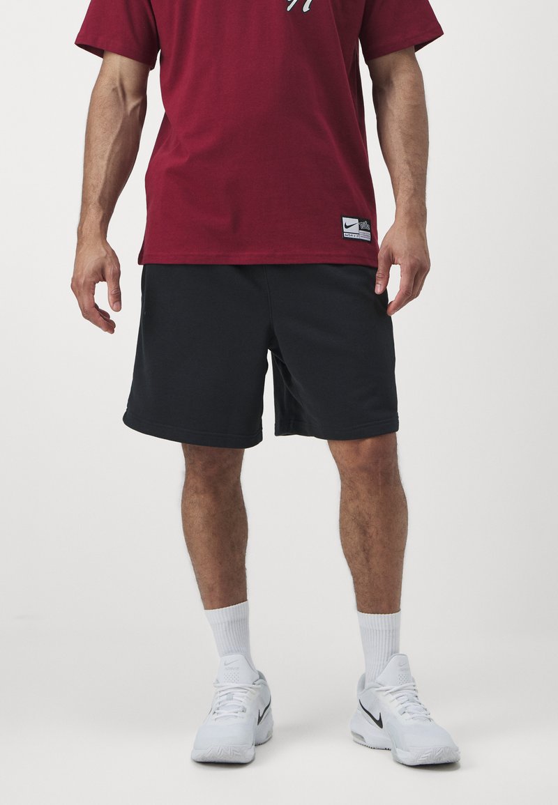 T-shirt vermelha de manga curta com logotipo, combinada com calções pretos e sapatilhas brancas, apresentando meias brancas até ao tornozelo. Design simples, ajuste casual.