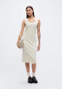 Ανοιχτό γκρι, αμάνικο φόρεμα από τζέρσεϊ με ίσια γραμμή και ένα μικρό λογότυπο στο στήθος. Το μοντέλο κρατά μια μπεζ τσάντα και φοράει μαύρα παπούτσια χωρίς κορδόνια.