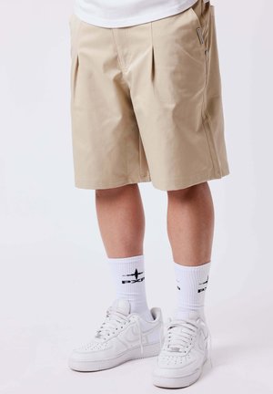 Man draagt beige knielange shorts, witte sportsokken met zwart logo en witte Nike-sneakers, staand tegen een witte achtergrond.
