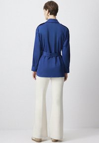 Donna in piedi di spalle con indosso una giacca blu a maniche lunghe con cintura e pantaloni bianchi a gamba larga, abbinati a scarpe beige.