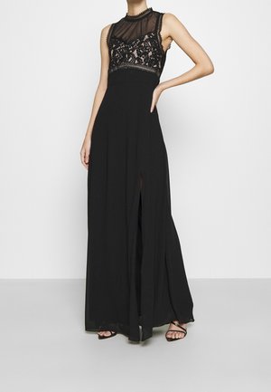 Vrouw die een zwarte mouwloze avondjurk draagt met een kanten lijfje en een doorschijnende halslijn, poseert met een hand op haar heup en draagt sandalen met bandjes.