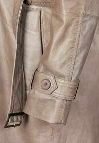 Helle beige Leder-Trenchcoat-Bündchen mit einem runden Knopf und verstellbarem Riemen, ausgestattet mit glatter Textur und präzisen Nähdetails.