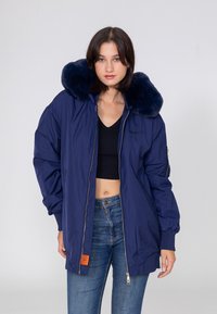 Marineblaue isolierte Jacke mit großem Kunstfellkragen, Reißverschluss vorne, elastischen Bündchen und orangefarbenem Etikett an dem unteren Saum.