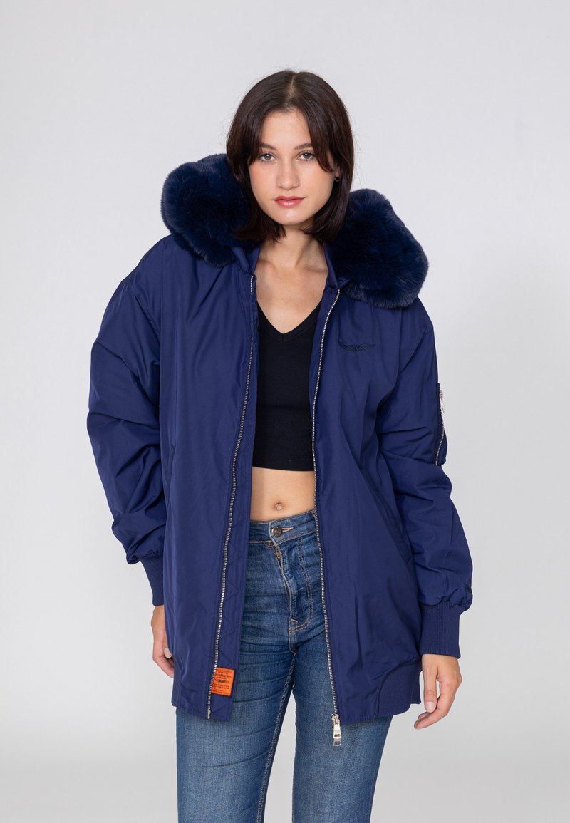 Marineblaue isolierte Jacke mit großem Kunstfellkragen, Reißverschluss vorne, elastischen Bündchen und orangefarbenem Etikett an dem unteren Saum.