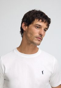 Biała bawełniana koszulka z okrągłym dekoltem i małym niebieskim haftem golfisty na lewej piersi. Gładka faktura, klasyczny krój.