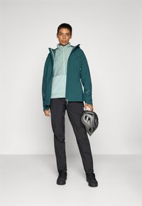 Jachetă de vreme rezistentă pentru femei, de culoare teal, cu un strat de bază verde deschis, asortată cu pantaloni negri și o cască gri, având fermoare și buzunare.