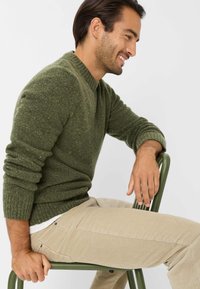 Grüner Strickpullover mit strukturiertem Stoff und gerippten Bündchen, kombiniert mit hellbeigen Cordhosen. Modell sitzt auf einem grünen Stuhl.