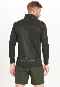Zwart sportshirt met lange mouwen, voorzien van een hoge kraag, mesh-panelen en een hartslaglijn-graphic op de achterkant, gecombineerd met olijfgroene shorts.