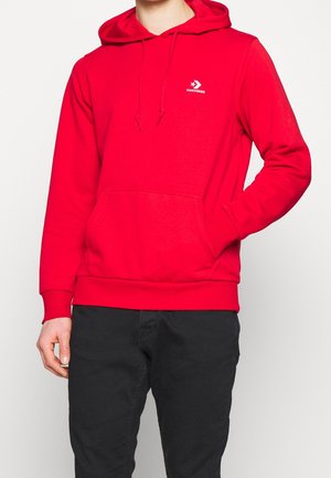 Hombre con sudadera con capucha roja brillante de Converse con bolsillo frontal y pantalones negros, posando contra un fondo liso.