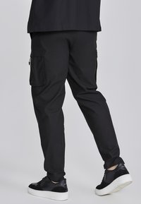 Pantalones cargo negros de corte slim, con bolsillos laterales, una textura suave y puños elásticos. Combinados con zapatos negros de suela blanca.