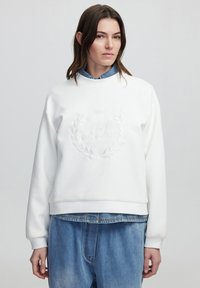 Sudadera blanca de cuello redondo con logo bordado y diseño de laurel. Presenta puños de canalé, corte relajado, llevada sobre una camisa de mezclilla y jeans holgados.