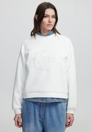Felpa bianca con collo a giro, dotata di logo ricamato e design a alloro. Presenta polsini a costine, vestibilità rilassata, indossata sopra una camicia di denim e jeans larghi.
