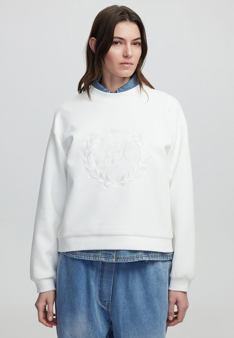 Sudadera blanca de cuello redondo con logo bordado y diseño de laurel. Presenta puños de canalé, corte relajado, llevada sobre una camisa de mezclilla y jeans holgados.