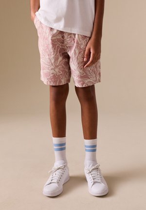 Barn iført lyserøde blomstrede shorts, hvid T-shirt, hvide sneakers og hvide sokker med to blå striber står på beige gulv.