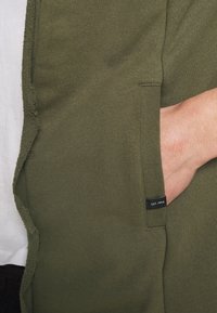 Sudadera verde oliva con un tejido de punto texturizado. Presenta bolsillos laterales y una pequeña etiqueta con "EST. 1993" cerca del dobladillo.