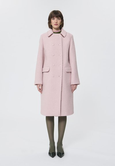 Manteau en laine rose à double boutonnage avec un col, deux poches avant et un tissu texturé, associé à des collants olive et des escarpins noirs à bout pointu.