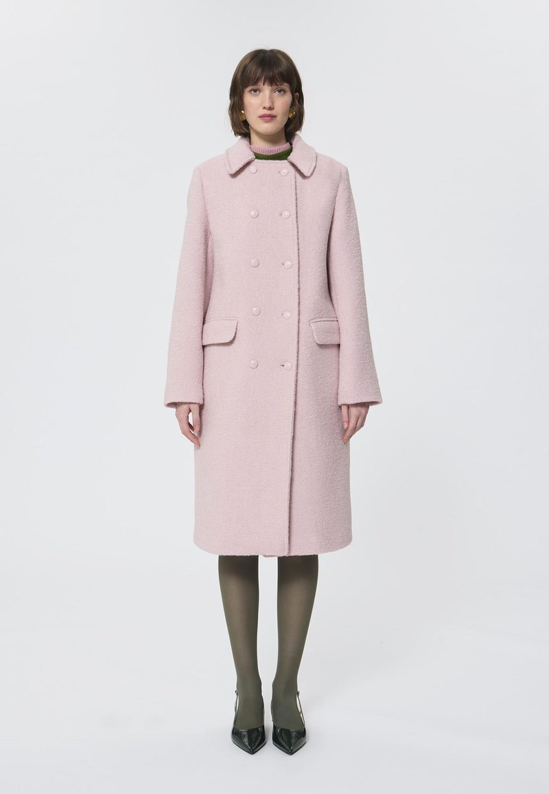 Cappotto rosa in lana doppiopetto con colletto, due tasche frontali e tessuto texturizzato, abbinato a collant oliva e décolleté nere a punta.