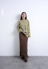 Femme posant contre un mur blanc, vêtue d'un haut à manches longues rayé vert olive et blanc, d'une jupe maxi marron et de chaussures marron foncé.