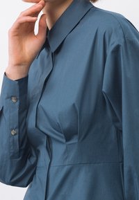 Blusa in cotone blu con colletto strutturato, polsini abbottonati e pieghe sottili lungo il davanti per un tocco di texture e dettaglio.