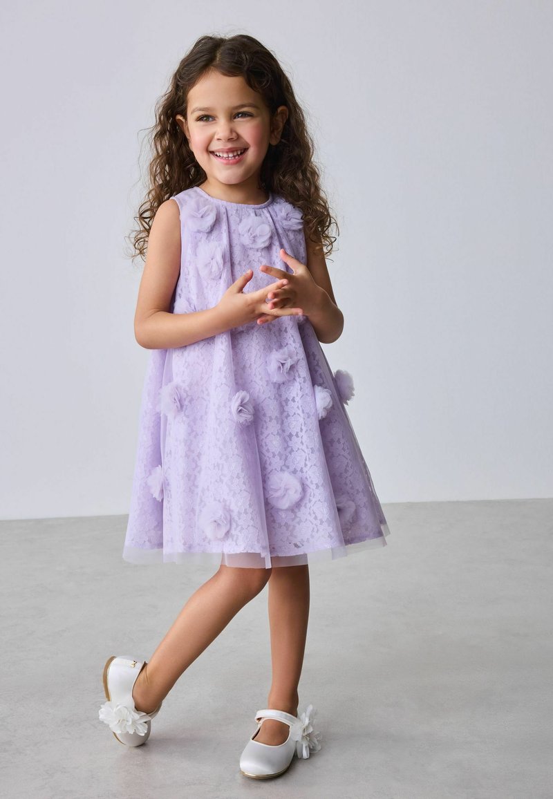 Usměvavá mladá dívka s kudrnatými vlasy, která má na sobě levandulové bezrukávé šaty s květinovými aplikacemi a bílé boty s květinovými detaily.