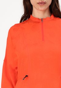 Heldere oranje zip-up top met een hoge kraag, voorzien van een subtiele gestructureerde textuur en een verstelbaar trekkoord in de taille.