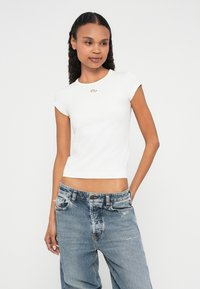 Vit figursydd t-shirt med korta ärmar och rund halsringning, med en liten logotyp på bröstet. Matchas med slitna blå jeans.