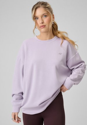 Młoda kobieta z blond kucykiem, ubrana w luźną lawendową bluzę i ciemne legginsy, stoi na jednolitym szarym tle.