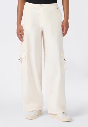 Femme portant un pantalon cargo ample couleur blanc cassé avec plusieurs poches et des baskets blanches, debout sur un fond uni.