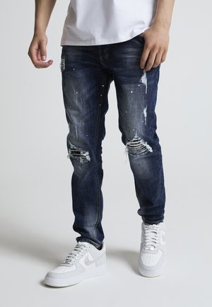 Personne portant un jean bleu foncé usé avec des déchirures et des éclaboussures de peinture blanche, associé à des baskets blanches, debout devant un fond uni.