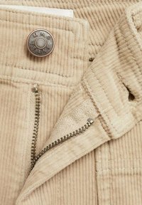 Beige corduroy-kangas, jossa on metallinen nappi, johon on merkitty "MNG JEANS," esillä oleva vetoketju ja tekstuurilliset ompeluyksityiskohdat.