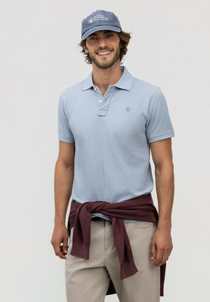 Hombre sonriente con una camisa polo azul claro, gorra azul marino, pantalones beige y un suéter granate atado alrededor de su cintura sobre un fondo liso.
