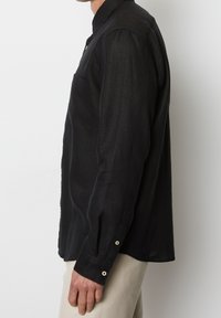 Camisa negra de manga larga con cuello abotonado, que cuenta con un bolsillo frontal y puños abotonados, hecha de una tela ligera y texturizada.
