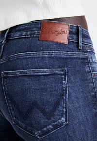 Jean en denim bleu foncé avec une étiquette en cuir marron Wrangler et un design classique de poches arrière, agrémenté d'un motif de surpiqûre décoratif subtil.
