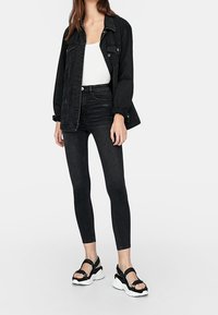 Chaqueta de denim negra sobre una blusa blanca, combinada con jeans ajustados negros y sandalias negras con suelas blancas. La tela tiene una textura suave.