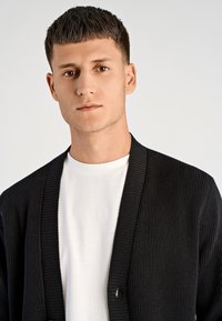 Czarny prążkowany kardigan z kołnierzem szalowym, otwartym przodem i jednym guzikiem, noszony na gładkiej białej koszulce z okrągłym dekoltem.
