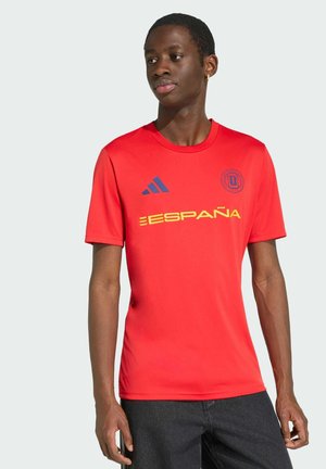 Ung mand iført en rød sportstrøje med teksten "España", Adidas-logoet og et rundt emblem, stående mod en ensfarvet lys baggrund.