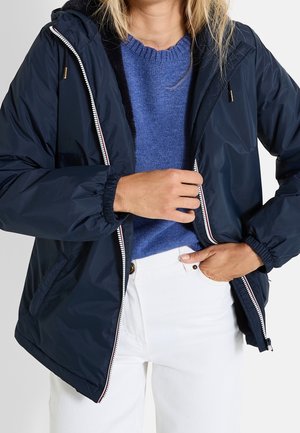 Mujer con una chaqueta azul marino sobre un suéter azul y pantalones blancos, con una mano ajustando la cremallera de la chaqueta y la otra en un bolsillo.