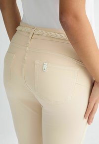 Pantaloni beji cu talie înaltă, având un brâu împletit, cu un singur buzunar posterior decorat cu un detaliu din metal argintiu. Textură netedă.