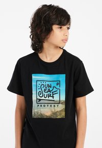Camiseta negra con un gráfico rectangular que muestra una escena de playa y texto estilizado "SUN SEA SURF PROTEST" en letras negras en negrita.