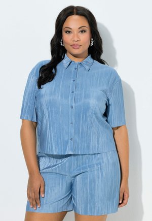 Frau mit langen dunklen Haaren trägt ein hellblaues, geripptes Kurzarm-Hemd mit Knöpfen und passende Shorts, steht vor einem einfarbigen hellen Hintergrund.