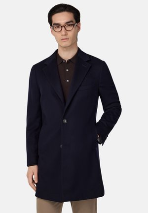 Navy wollen jas met een getailleerde pasvorm, enkelbreasted ontwerp, gekleurde revers, twee knopen en een linker borstzak. Draagt een bruine polo.