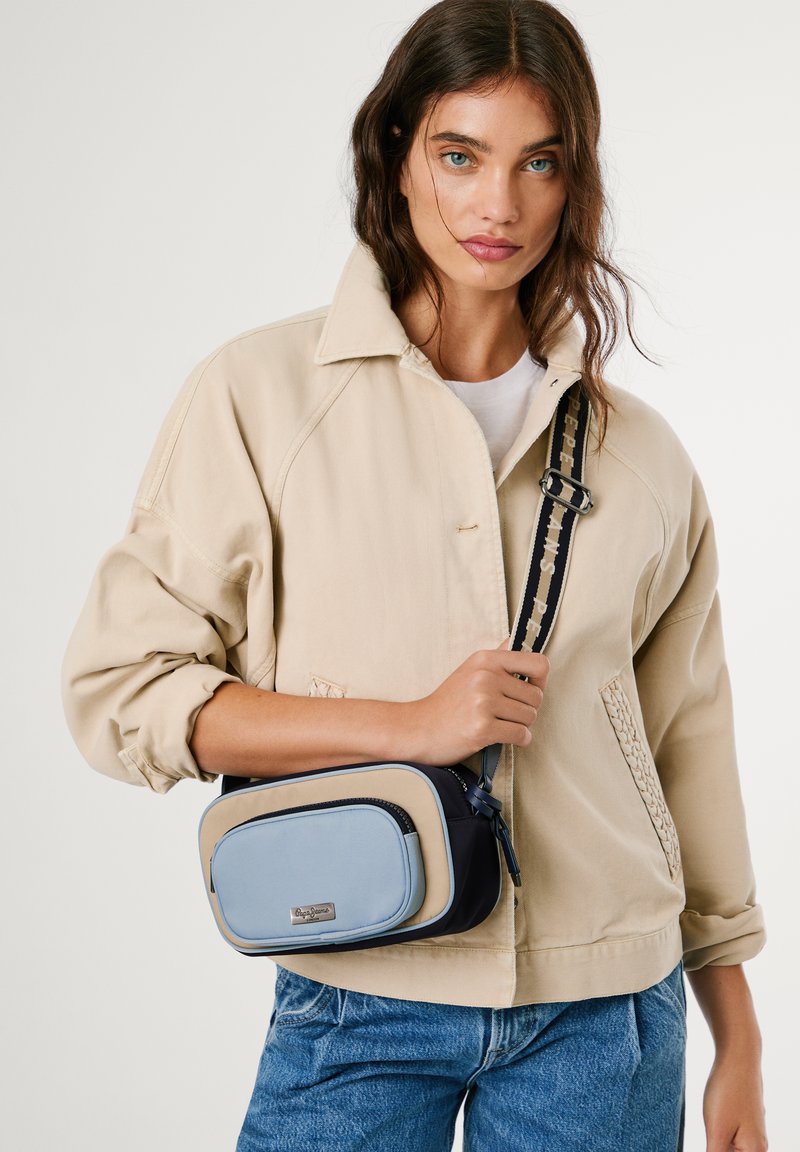 Kvinna som bär en beige jacka och blå jeans, håller en blå och beige axelväska med justerbar rem.