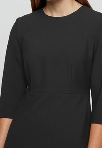 Svart klänning med rund halsringning och trekvartslånga ärmar. Har en texturerad tyg och subtil sömdetaljering vid midjan.