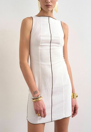 Vrouw draagt een mouwloze witte jurk met verticale zwarte naden, grote gouden en zilveren armbanden en gele geometrische oorbellen.