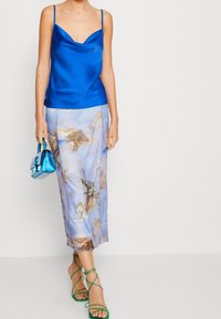 Blå satin camisole med en cowl-halsringning, parat med en ljusblå kjol som har klassiska konsttryck. Gröna sandaler med remmar och en liten blå väska.