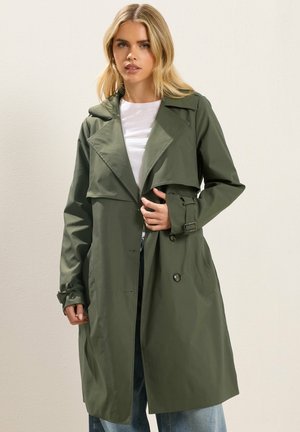 Femme portant un long trench-coat vert olive avec des détails à la ceinture, debout devant un fond clair uni, ajustant l'avant du manteau.
