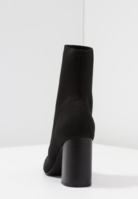 Bottine noire avec un dessus en textile extensible, talon bloc haut, texture lisse et design minimaliste. Vue arrière montrant les détails de couture.