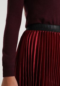 Bordeaux trui gecombineerd met een rode geplooide rok. De rok heeft een zwarte ribbelband in de taille en de stof lijkt glanzend en soepel te zijn.