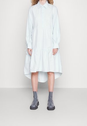 Femme portant une robe-chemise bleu clair à manches longues avec un ourlet asymétrique et des bottines grises brillantes, debout devant un fond uni.